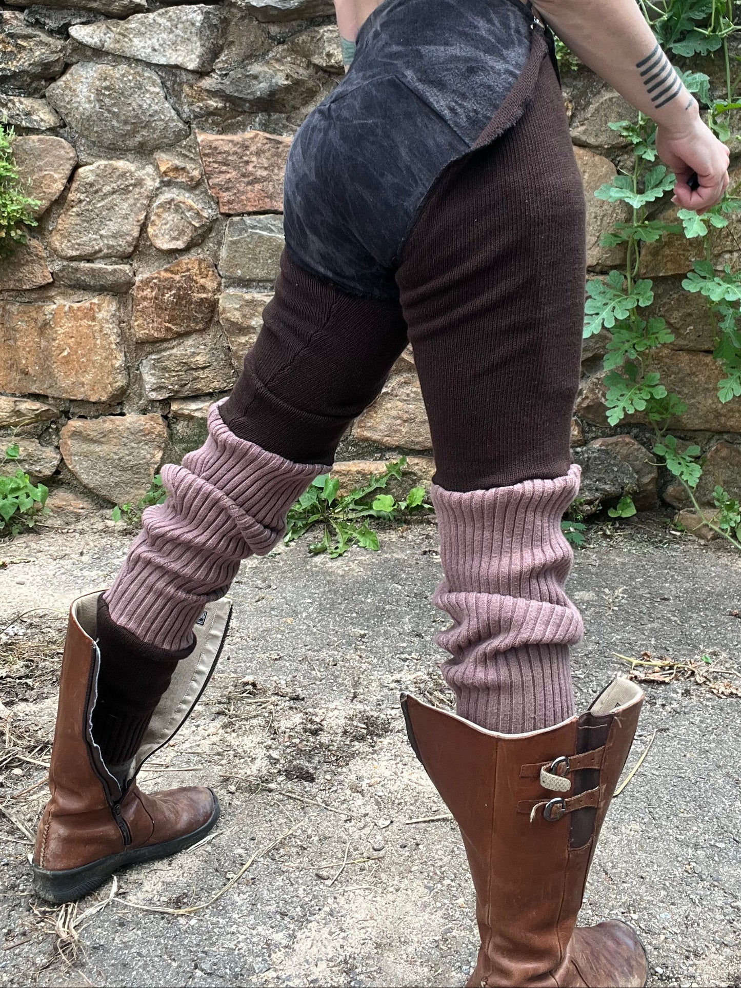 A person outdoors wearing a dark brown and black patchwork apron-style top, dark brown corduroy pants, mauve ribbed leg warmers, and brown leather boots, standing in front of a stone wall.