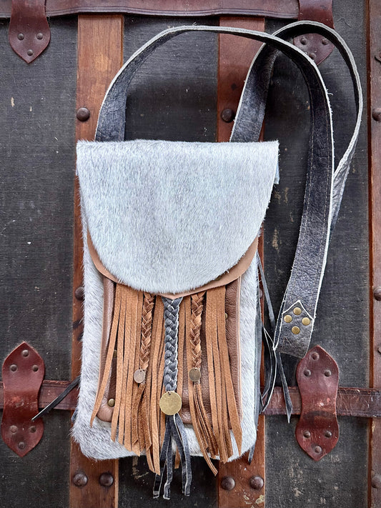 A white and grey fur cross-body purse with a short rounded lid, featuring hand cut fringe, leather braids, and antiqued brass hardware, displayed on a wooden surface with metal rivets.