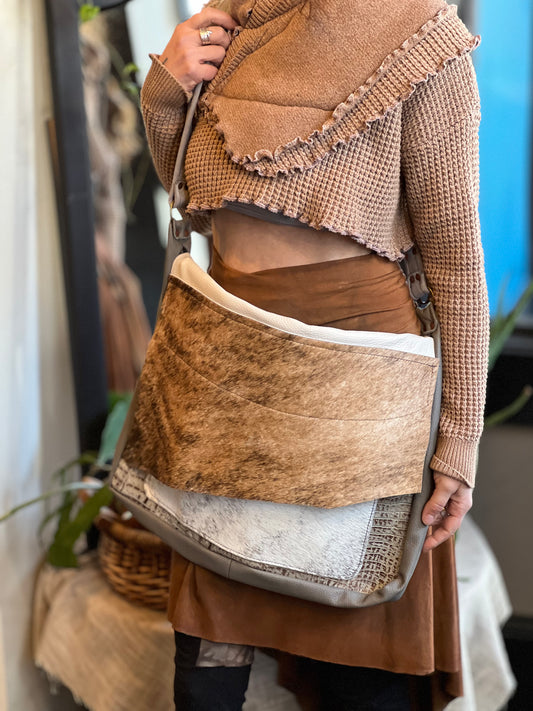 A person holding a brown fur and gator messenger bag with a shoulder strap and zippered pockets.