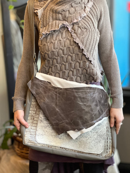A person wearing a grey sweater is carrying a brown leather messenger bag with a shoulder strap. The bag appears to be empty and is displayed in a vertical position.