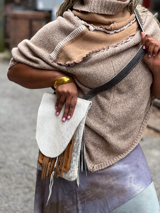 A person wearing a beige and gray leather cross-body purse with a fringe detail and antique brass hardware.