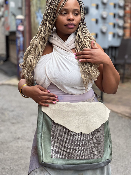 A person wearing a white shirt is holding a green and beige leather messenger bag with a shoulder strap.