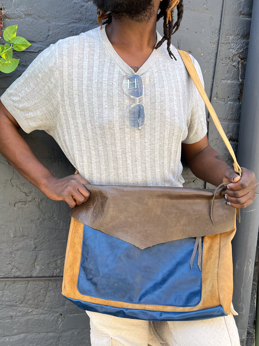 A person wearing a beige sweater is holding a cobalt blue and chestnut brown messenger bag across their body.