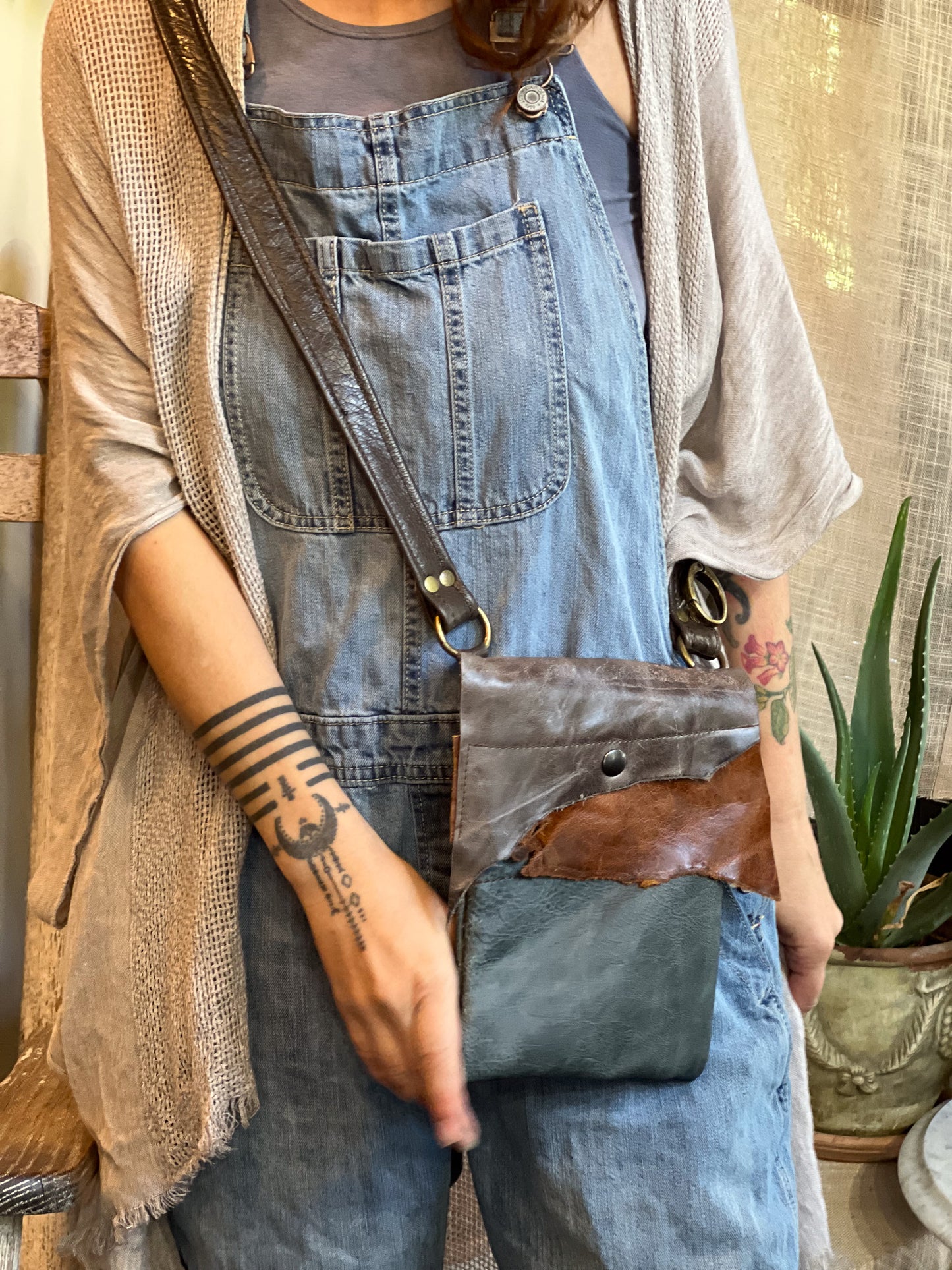 Person wearing denim overalls holding a crossbody bag with a plant in the background