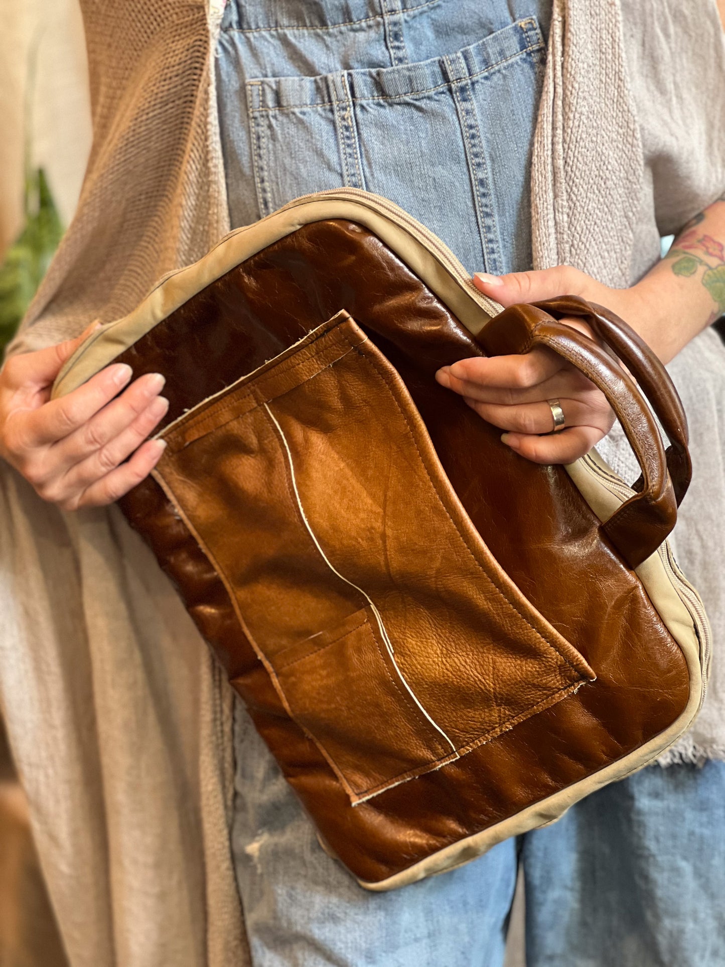 Brown leather bag held by a person wearing a beige cardigan and blue jeans.
