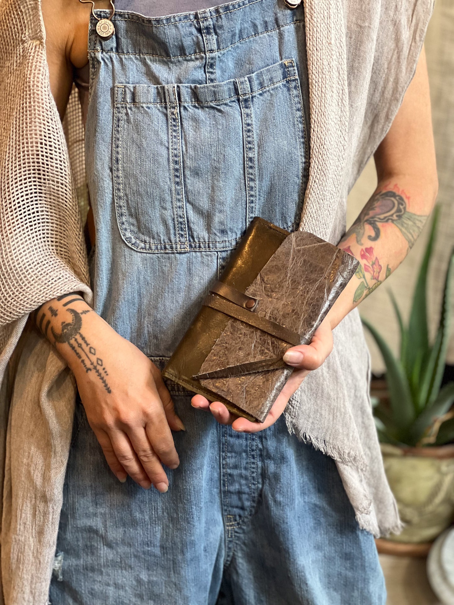 Person holding a brown leather wallet with tattoos visible on arms, wearing denim overalls.