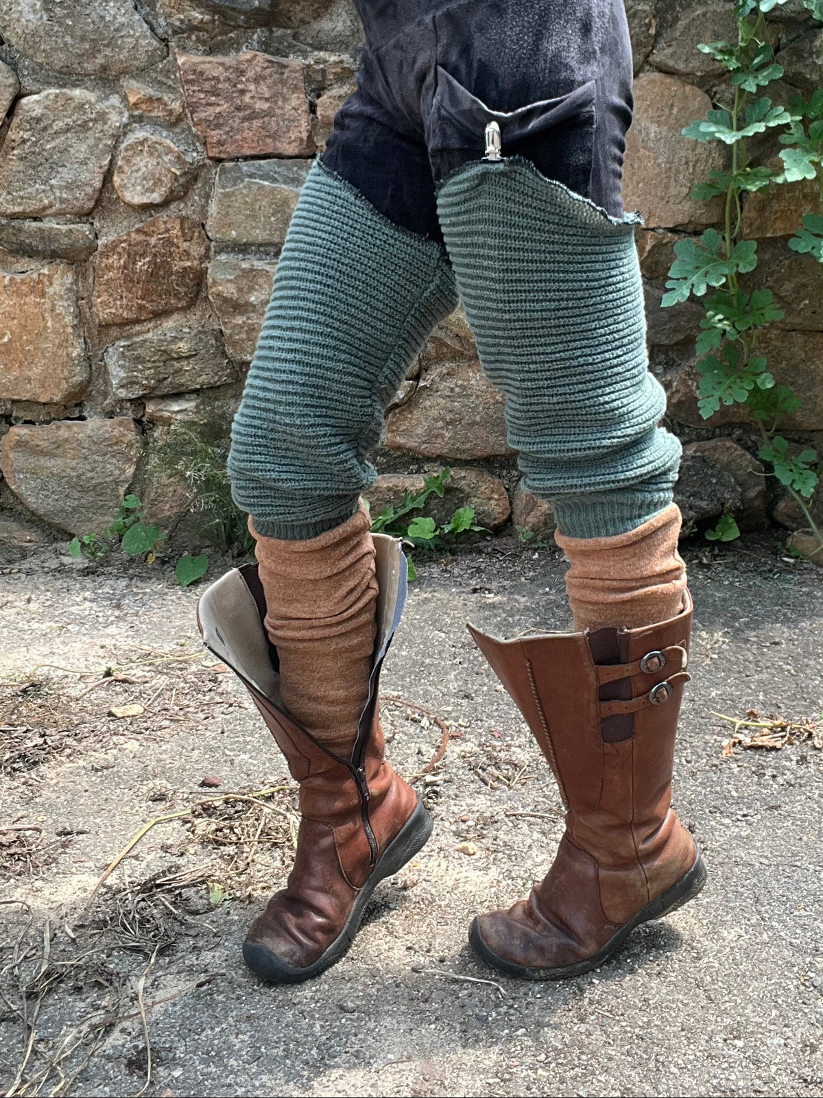 A person outdoors wearing a dark gray and black patchwork skirt with ribbed green leg warmers and brown leather knee-high boots, standing in front of a stone wall and greenery.