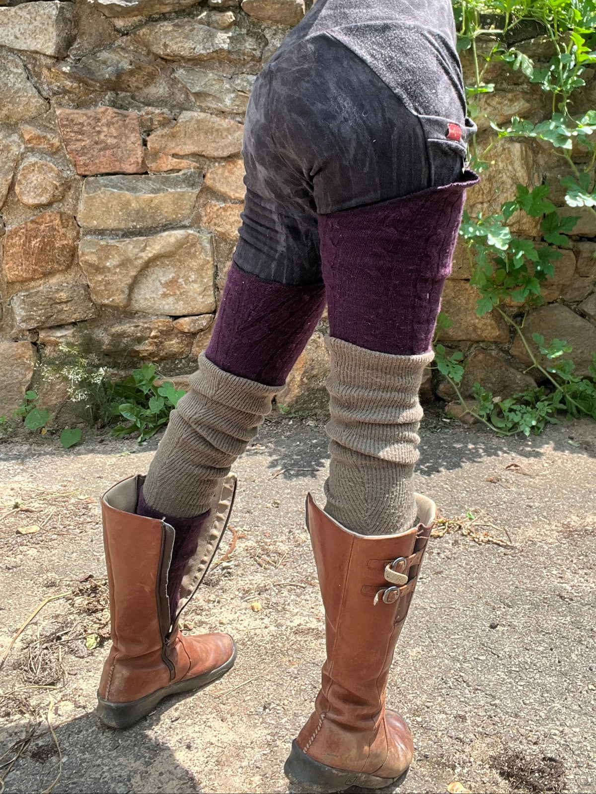 A person outdoors wearing dark gray patchwork shorts, purple knit leg warmers, beige knit boot covers, and brown leather boots, standing in front of a stone wall and greenery.