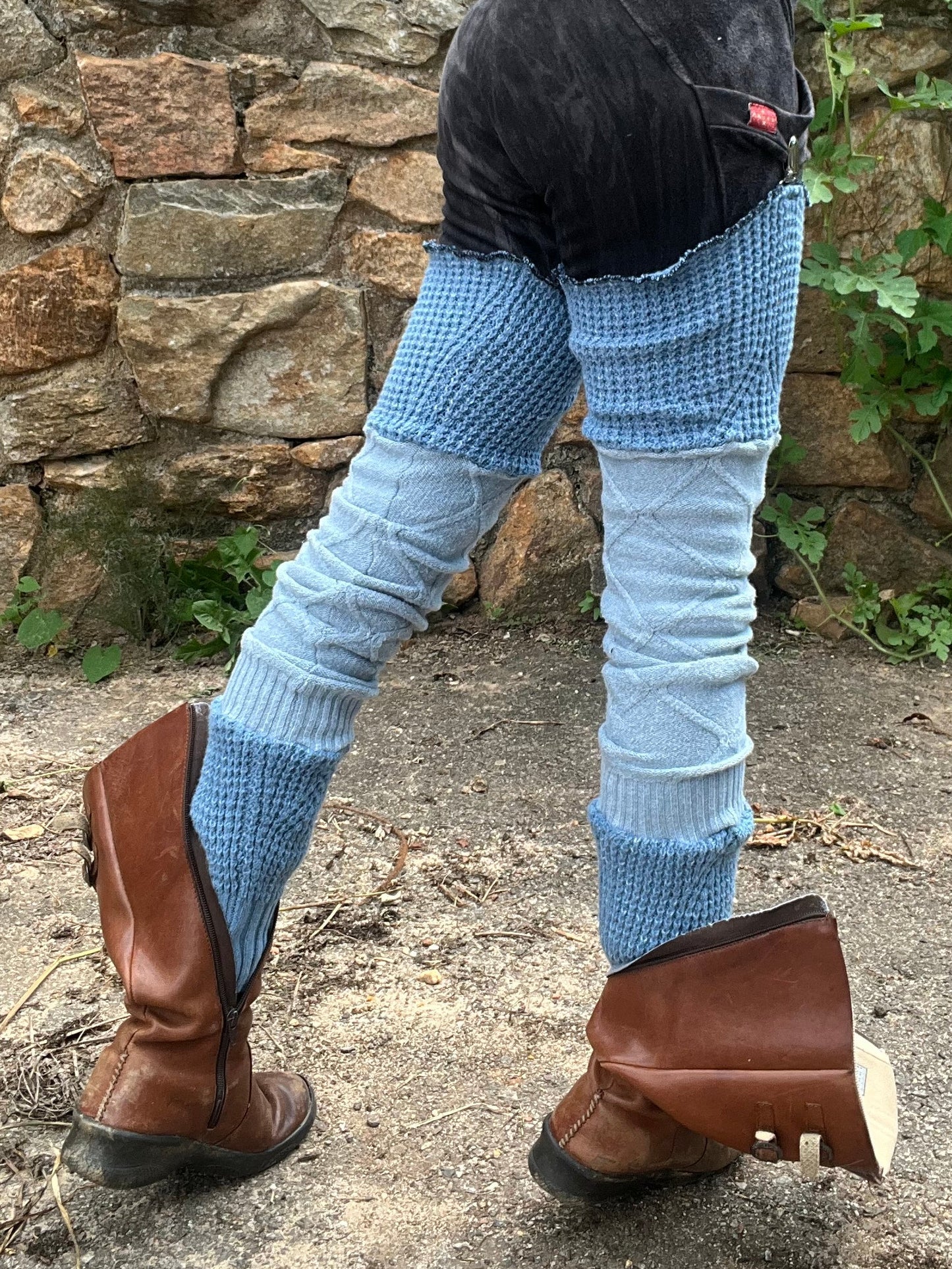 A person outdoors wearing a maroon cropped tank top, black shorts, light blue patterned leg warmers, and brown leather boots, standing in front of a stone wall and greenery.