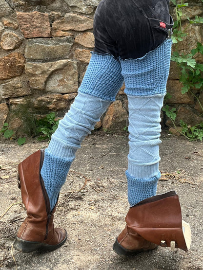 A person outdoors wearing a maroon cropped tank top, black shorts, light blue patterned leg warmers, and brown leather boots, standing in front of a stone wall and greenery.