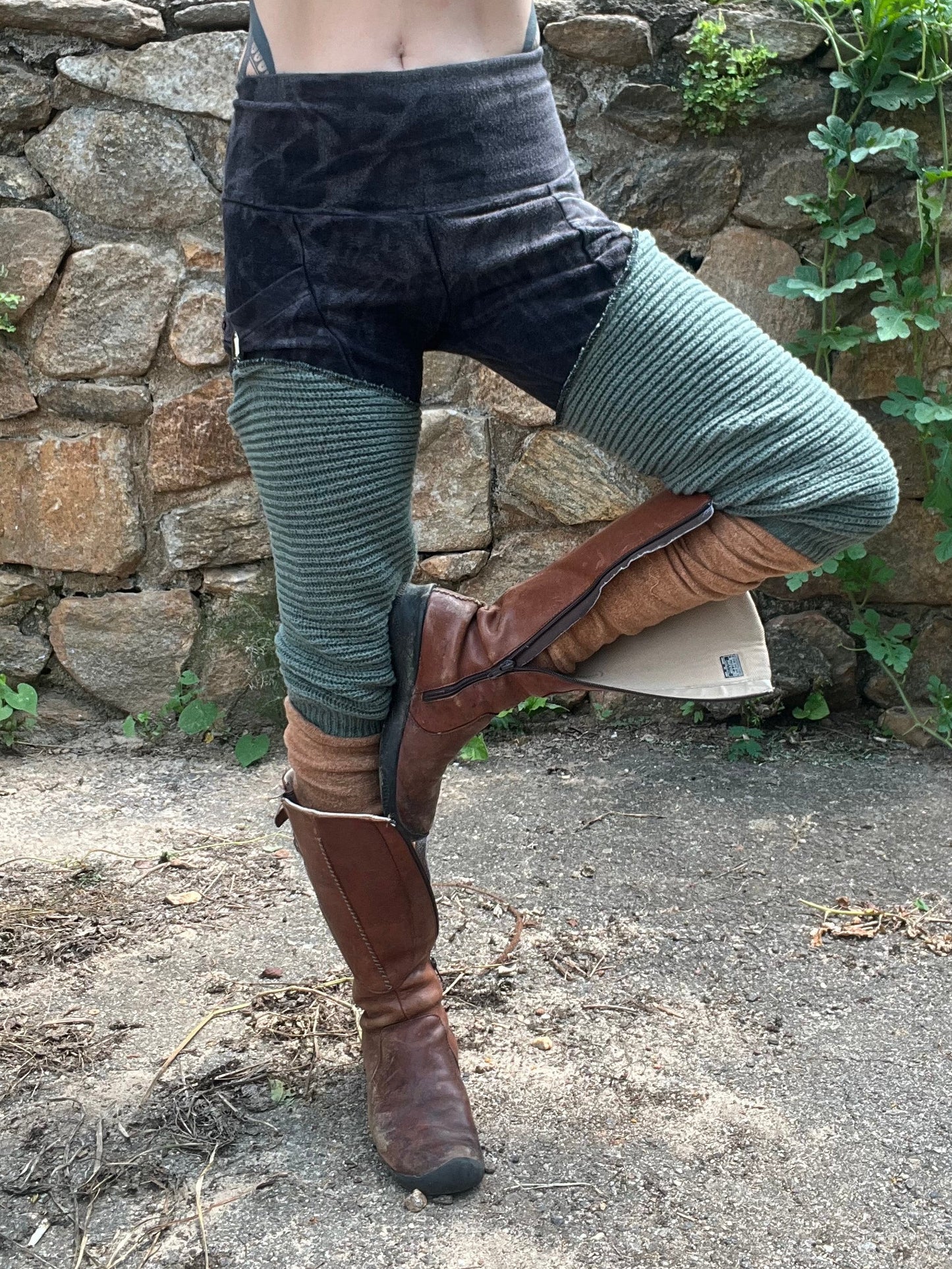 A person outdoors wearing a purple ribbed crop top, black and green ribbed leggings, and brown knee-high boots, standing in front of a stone wall with greenery.