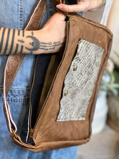 Brown leather bag with textured panel held by a person wearing a blue denim jacket.