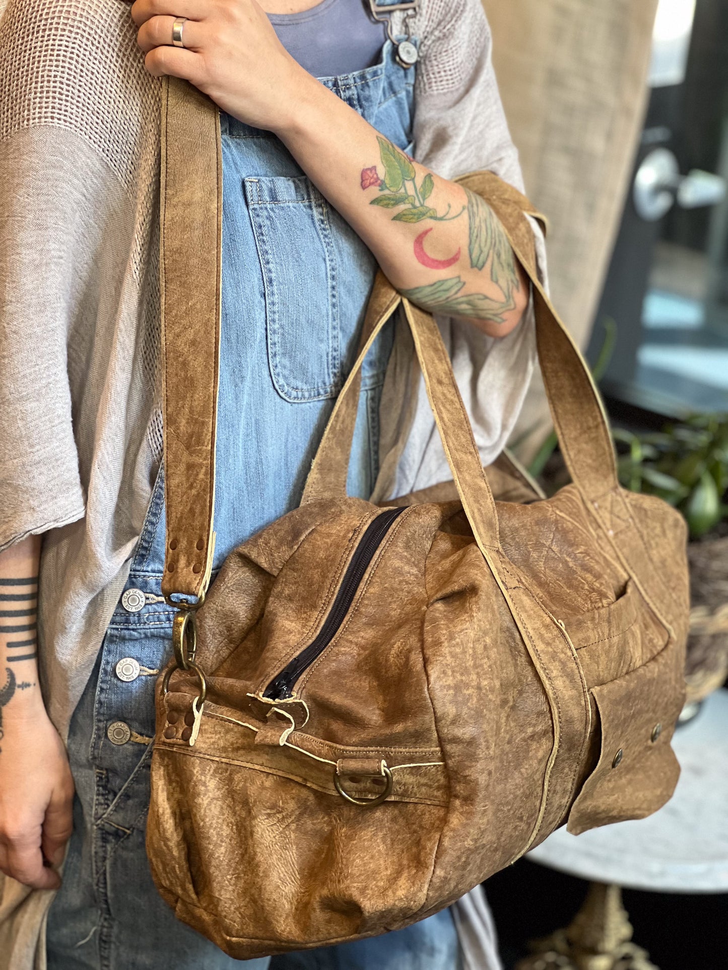 Person holding a brown leather bag with tattoos visible on arms