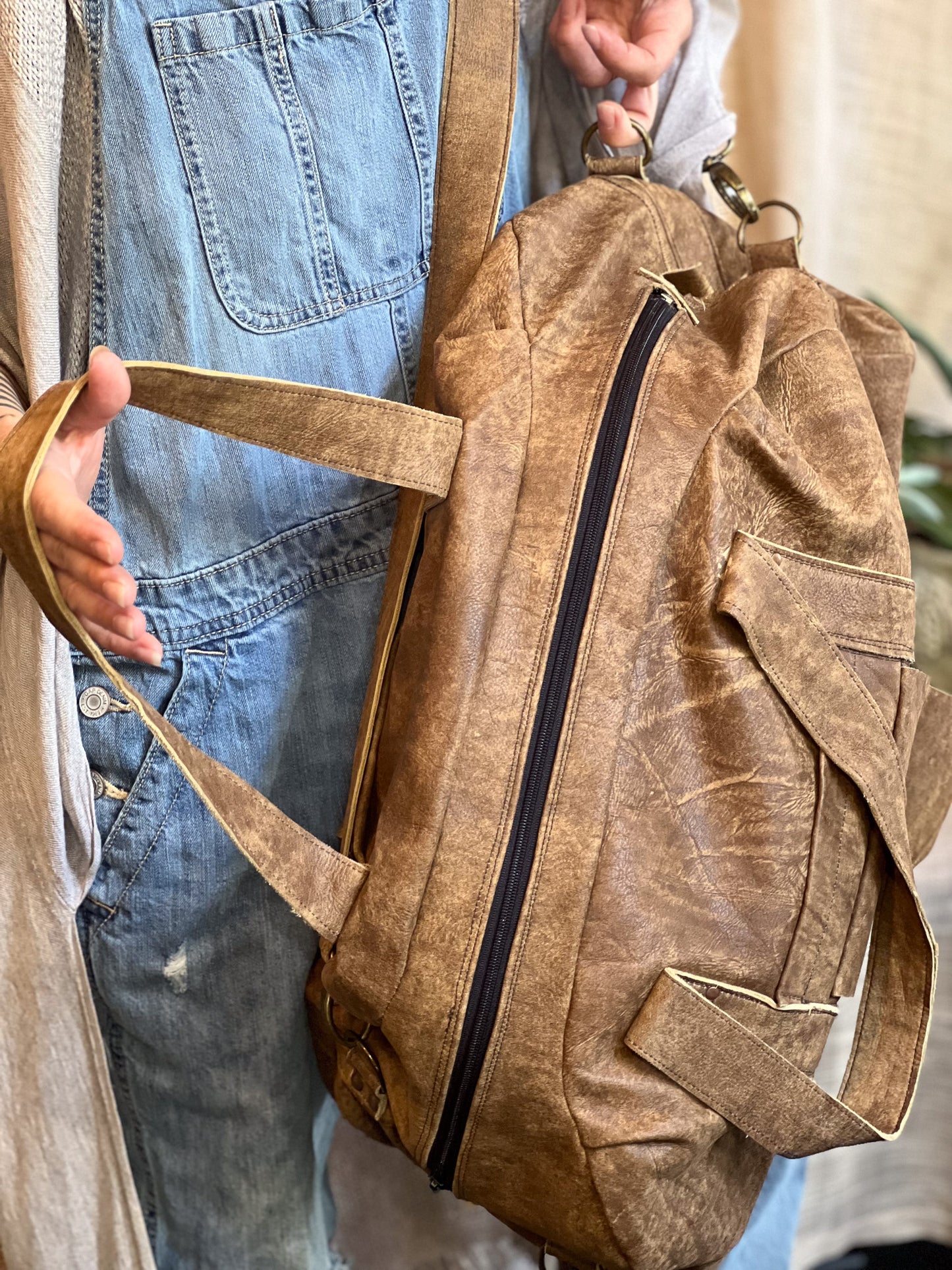 Brown leather backpack held by a person wearing blue overalls.