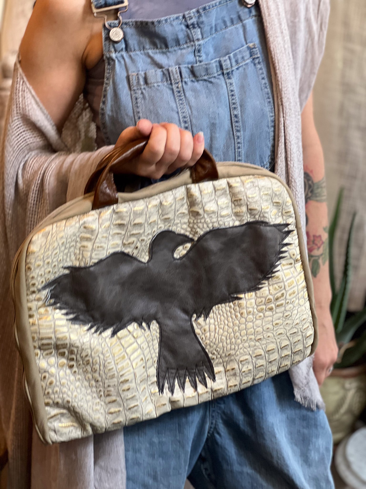 Person holding a beige handbag with a black bird design, wearing blue overalls.