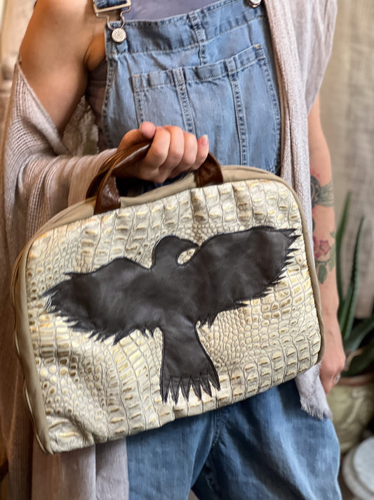 Person holding a beige handbag with a black bird design, wearing blue overalls.