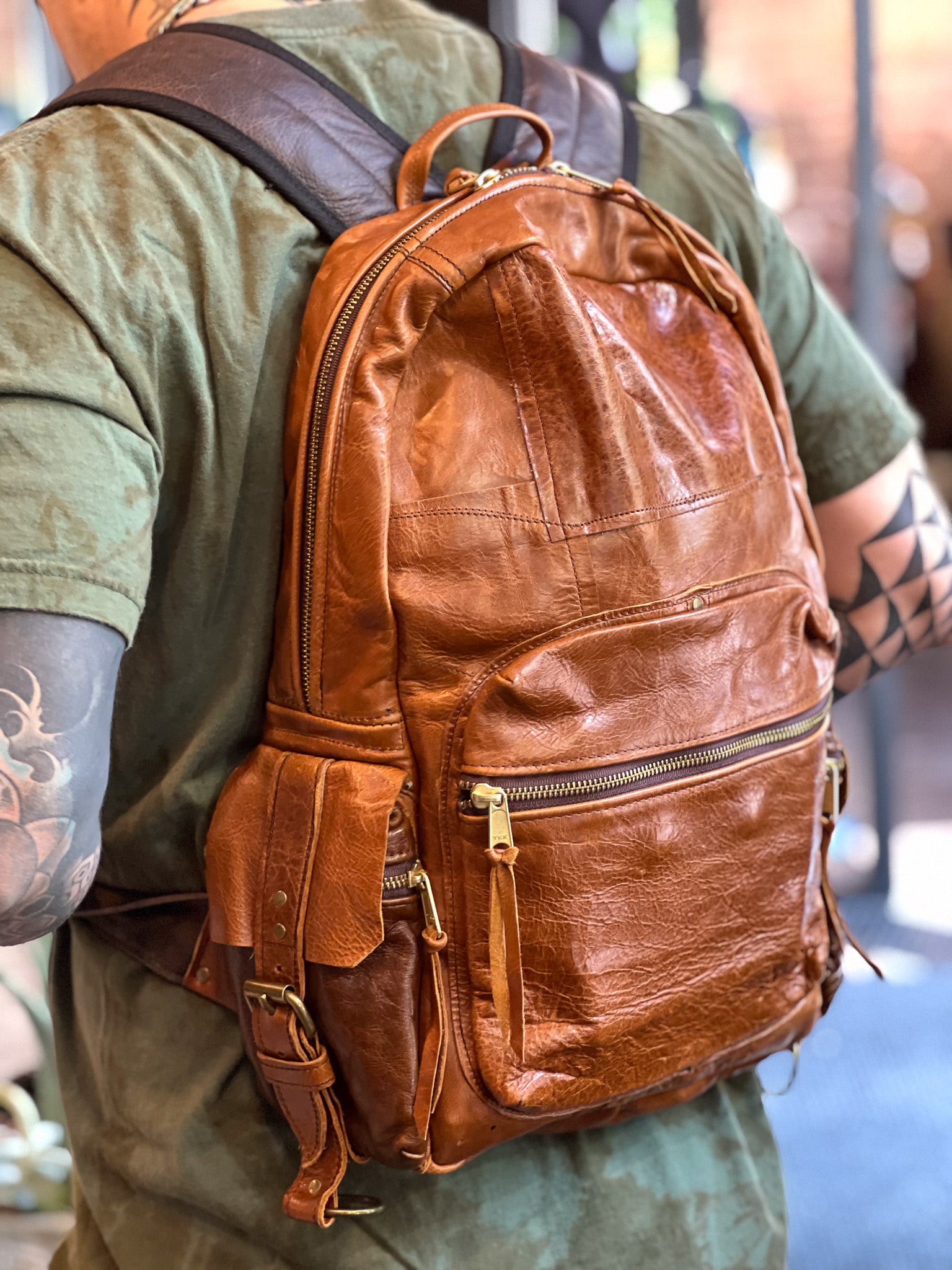 Brown leather backpack worn by a person with tattoos on a blurred background