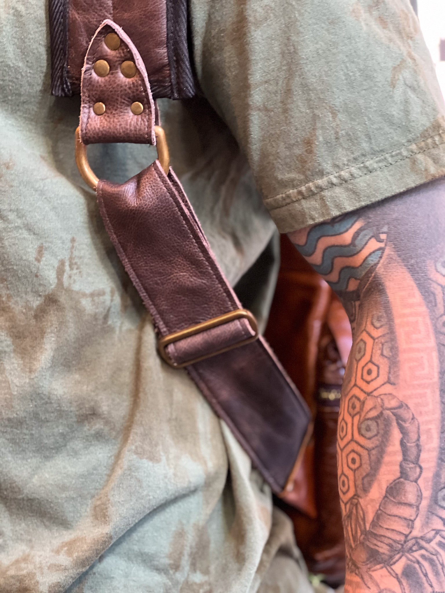 Brown leather strap with gold hardware on a person wearing a beige shirt and having a tattoo on their arm.