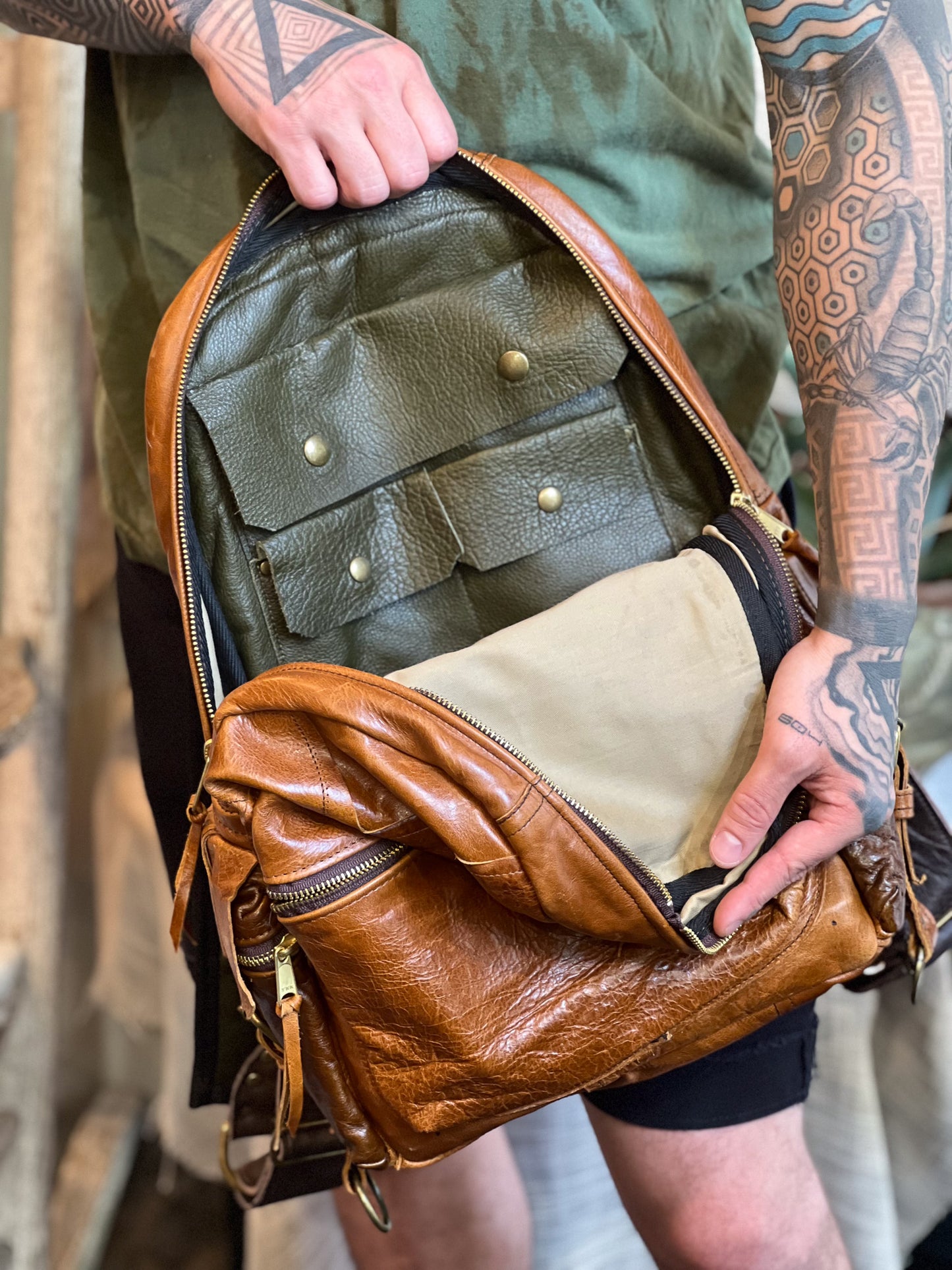 Brown leather backpack with multiple pockets held open by a person with tattoos.