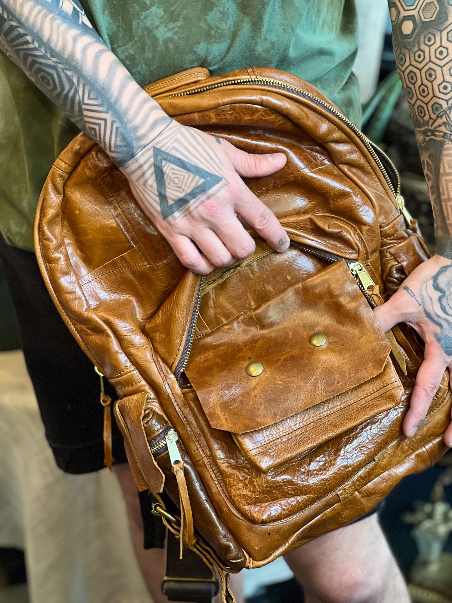 Brown leather backpack held by a person with tattoos on their arms.