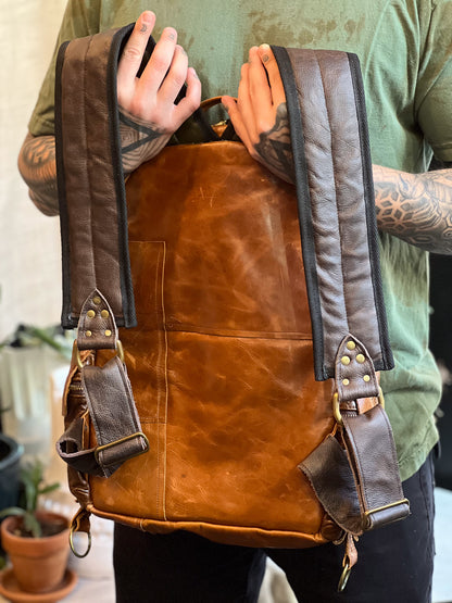 Brown leather backpack held by a person with tattoos, wearing a green shirt.