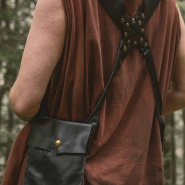 A woman model showcasing the back of the black leather pocket holster; showing of the 'X' strap in the back and how it sits on the shoulders. One of the two big pockets are shown, closed, with its brass snap closure.