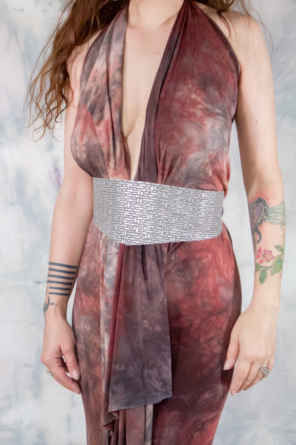 A woman model showcasing a silver leather, wrap-around cincher belt that has a futuristic, embossed pattern on the front; wearing it in the middle of her torso, cinching in the dress she is wearing.