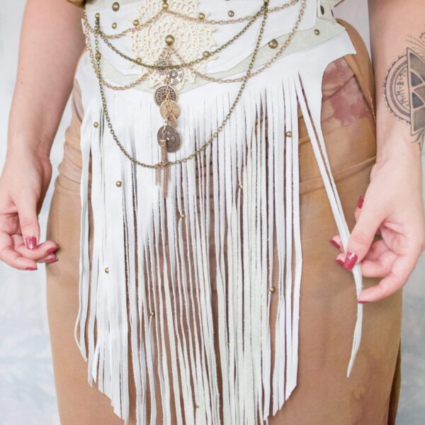 A woman model showcasing a white leather, long fringe, wrap-around cincher belt that has draped chains, antique studs, and other embellishments on it for decoration. She is wearing it in the middle of her torso, cinching in her dress.