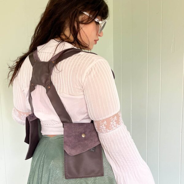 A woman model showcasing the back of the purple leather pocket holster; showing of the 'X' strap in the back and how it sits on the shoulders. One of the two big pockets are shown, closed, with its brass snap closure.