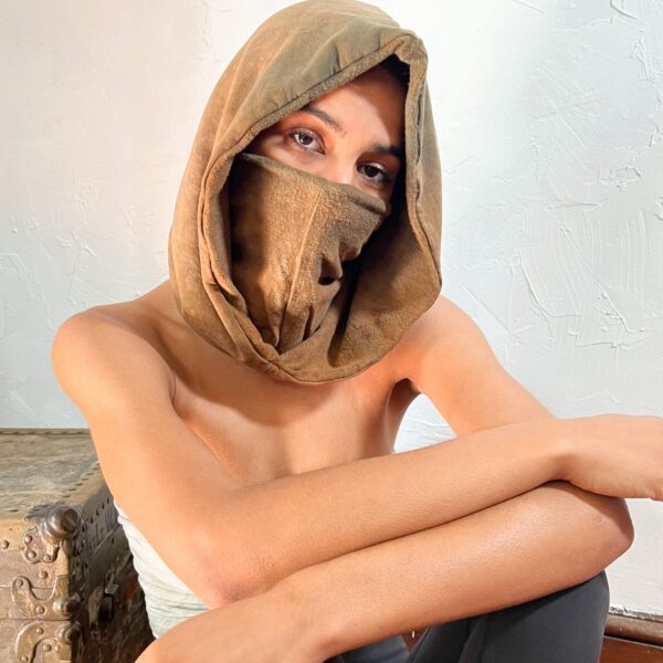 A woman model showcasing a unisex tan-colored, bamboo fleece, standalone hood; She is wearing the hood and has the entrance to the hood turned inward, and pulled up over her face like a mask.