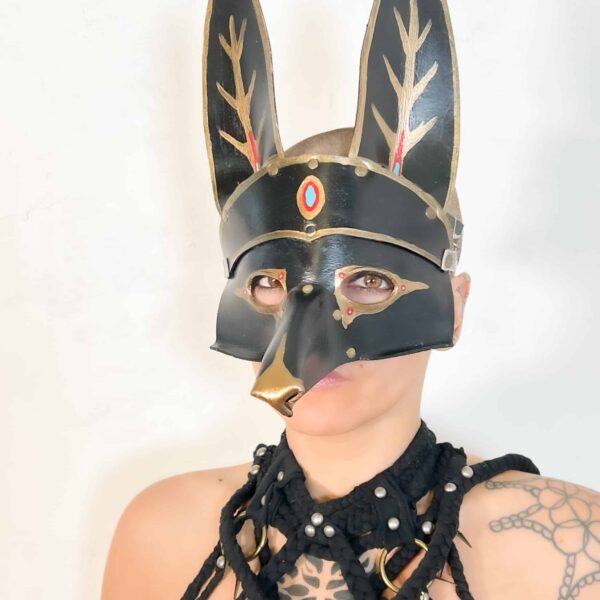 A woman model showcasing a handmade, black leather Anubis mask, that has painted gold, red & blue accents, and a snout that extends about 5 inches off the mask. 2 ears stand about 0 inches tall on the top part of the mask.