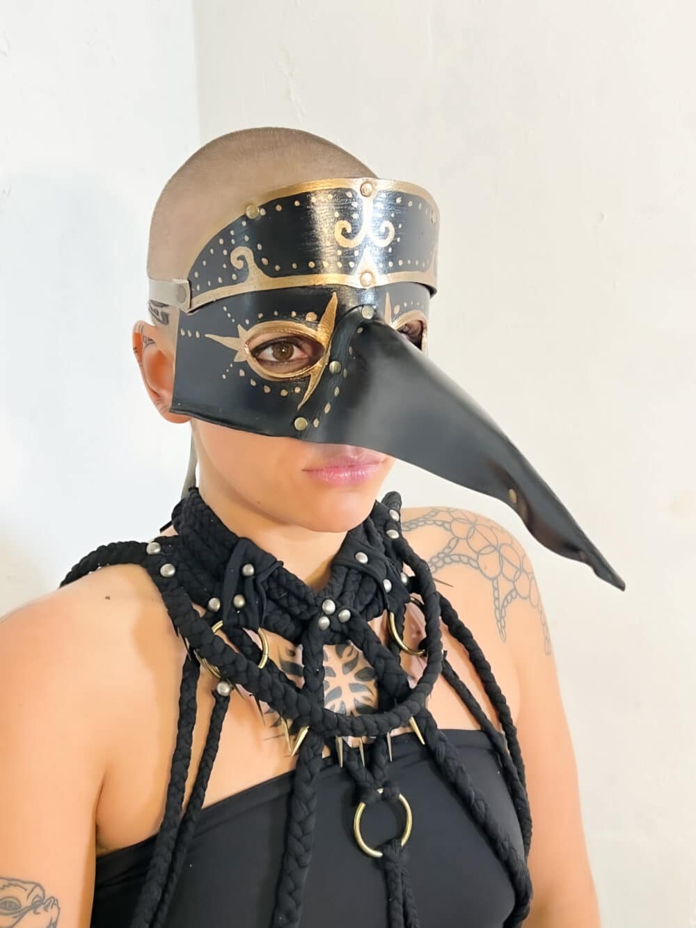 A woman model showcasing a handmade, black leather mask, that has a large beak that extends about 8 inches from the middle of the mask. This mask features eye holes, as well as gold-painted accents; a tan leather strap can be seen on the side wrapping around the head: this is the straps that tie together to hold the mask on the face.