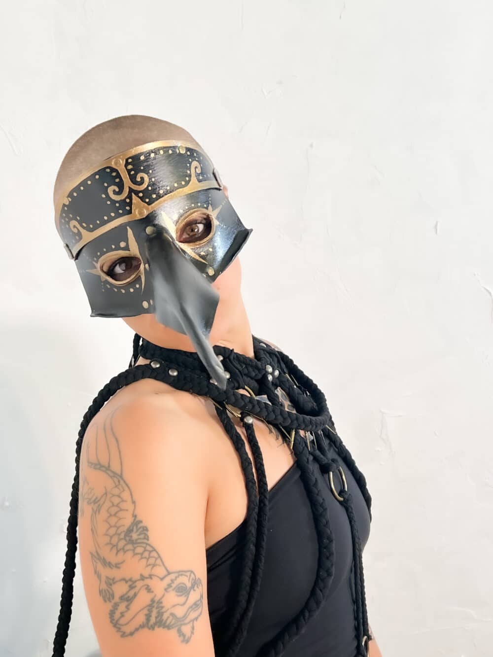 A woman model showcasing a handmade, black leather mask, that has a large beak that extends about 8 inches from the middle of the mask. This mask features eye holes, as well as gold-painted accents.