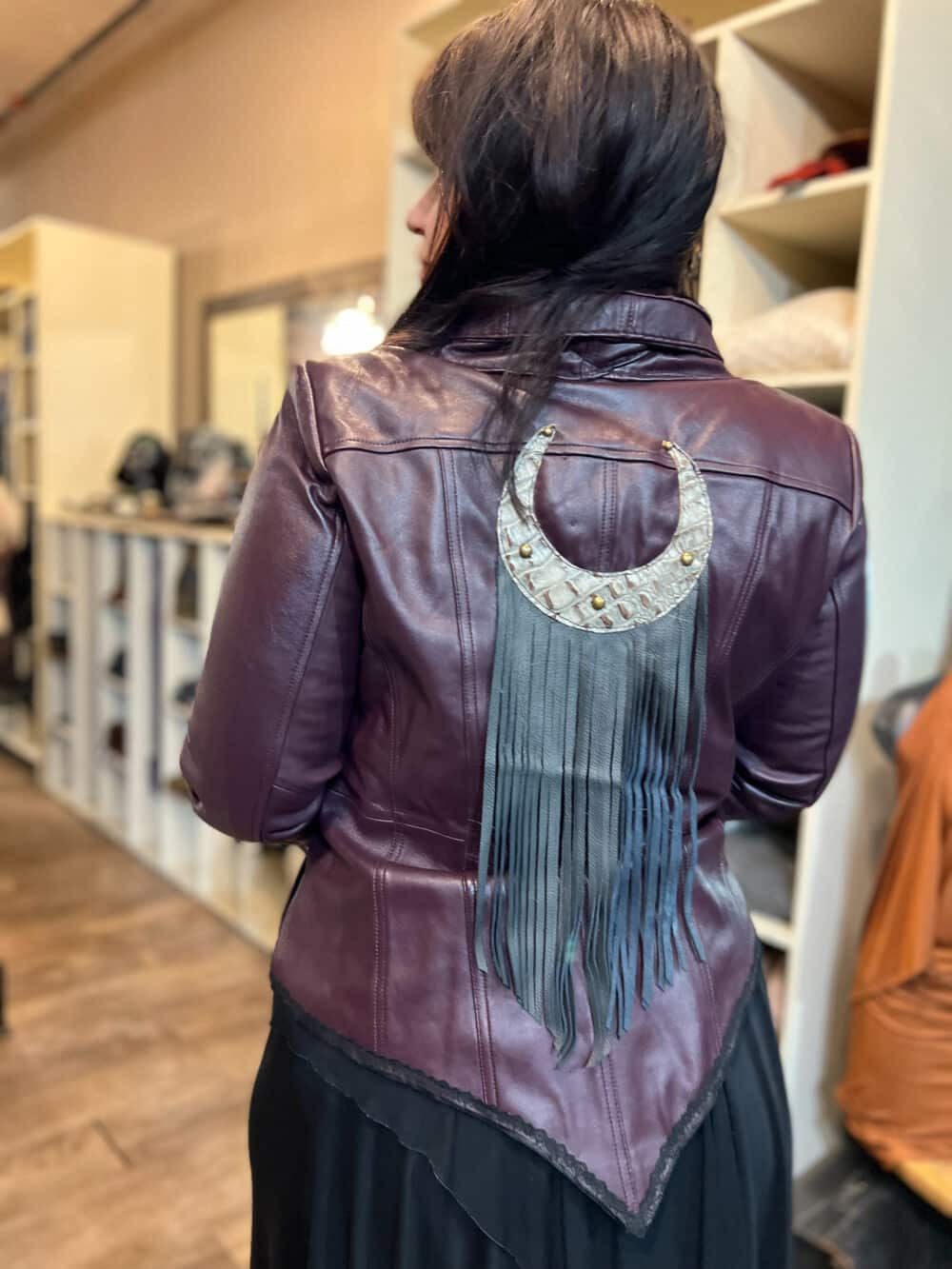 The back of a burgundy leather motorcycle jacket. A textured lunar moon emblem with long black fringe attached to the bottom takes up the back of the jacket.